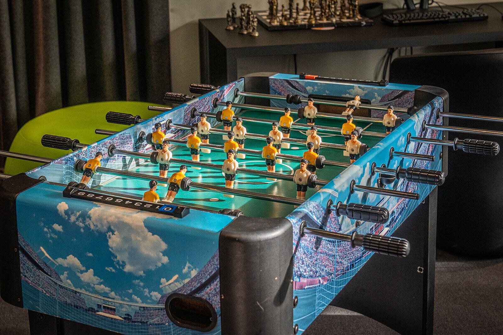 A foosball table with a blue and green color scheme, located in a room with a desk and keyboard in the background.