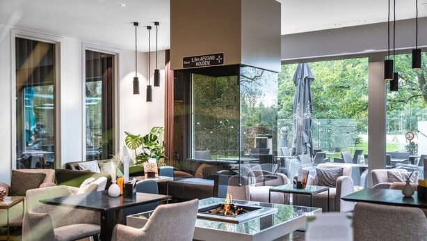 A modern indoor seating area with a central fire feature, large windows, and a view of the outdoors. - Velotel Brugge (Bruges)