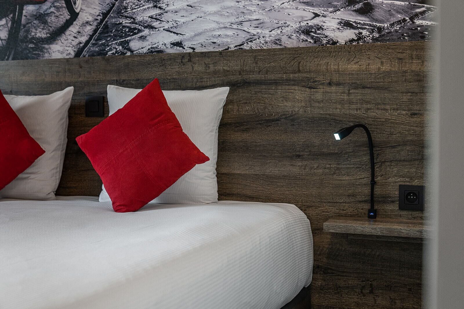 A bed with white bedding and red pillows, a wooden headboard, a bedside table with a lamp, and a wall with a snowy landscape mural.