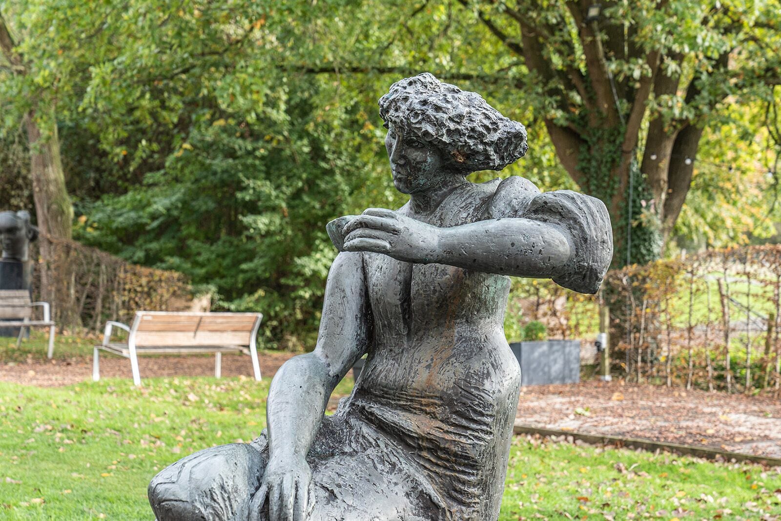 Eine Statue einer sitzenden Frau in einem Park, im Hintergrund eine Bank.