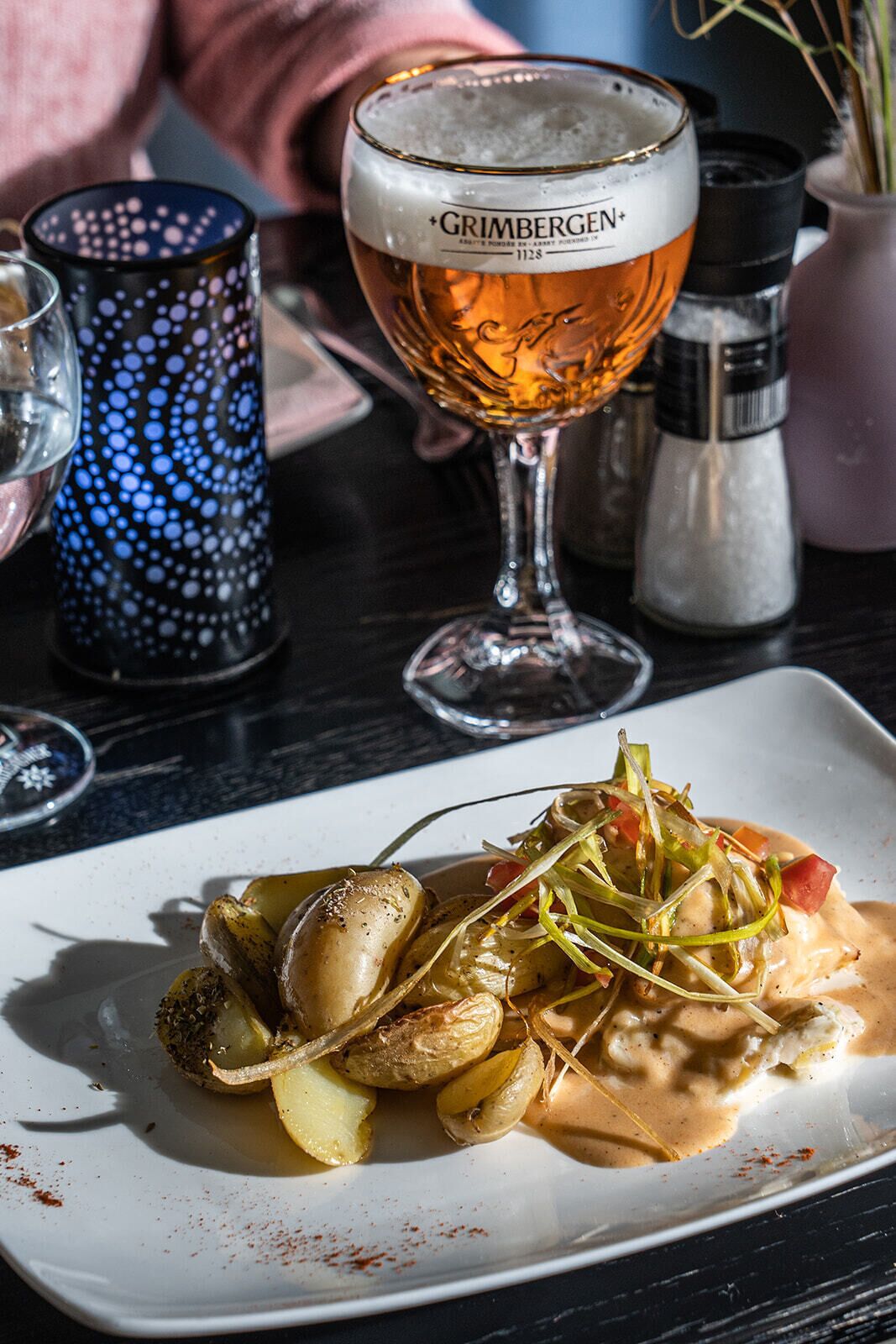 Een bord geroosterde aardappelen met een romige saus, een glas bier en een decoratieve kandelaar op een tafel.