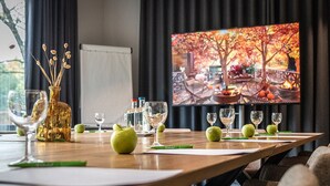 A conference room with a long table set for a meeting, a television displaying a scenic autumn landscape, and a decorative bottle on the table. - Velotel Brugge (Bruges)