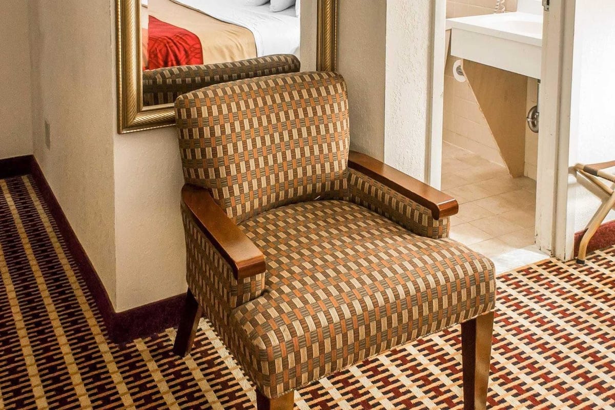 Une chambre d’hôtel avec un fauteuil en osier, un grand miroir, un lit avec du linge de lit blanc et une salle de bain avec un lavabo blanc.