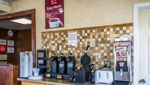 Espaço de café da manhã com máquina de waffles, cafeteiras e torradeira. Um aviso indica o horário do café da manhã e um cartaz divulga 'Easy Starts'.