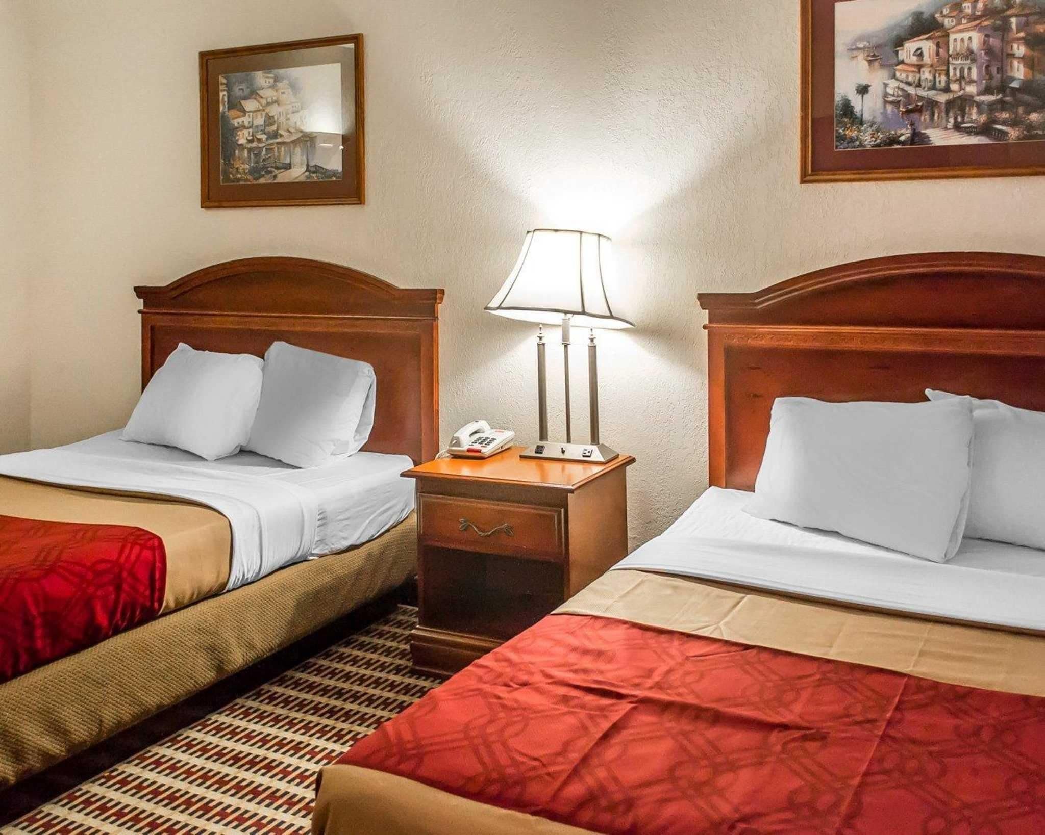Habitación de hotel con dos camas, cada una con cabecero de madera, ropa de cama blanca y una manta roja. Entre las camas, hay una mesita de noche con una lámpara y un teléfono.