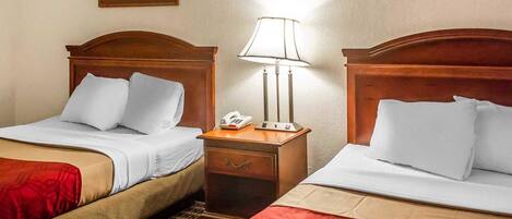 Habitación de hotel con dos camas, cada una con cabecero de madera, ropa de cama blanca y una manta roja. Entre las camas, hay una mesita de noche con una lámpara y un teléfono.