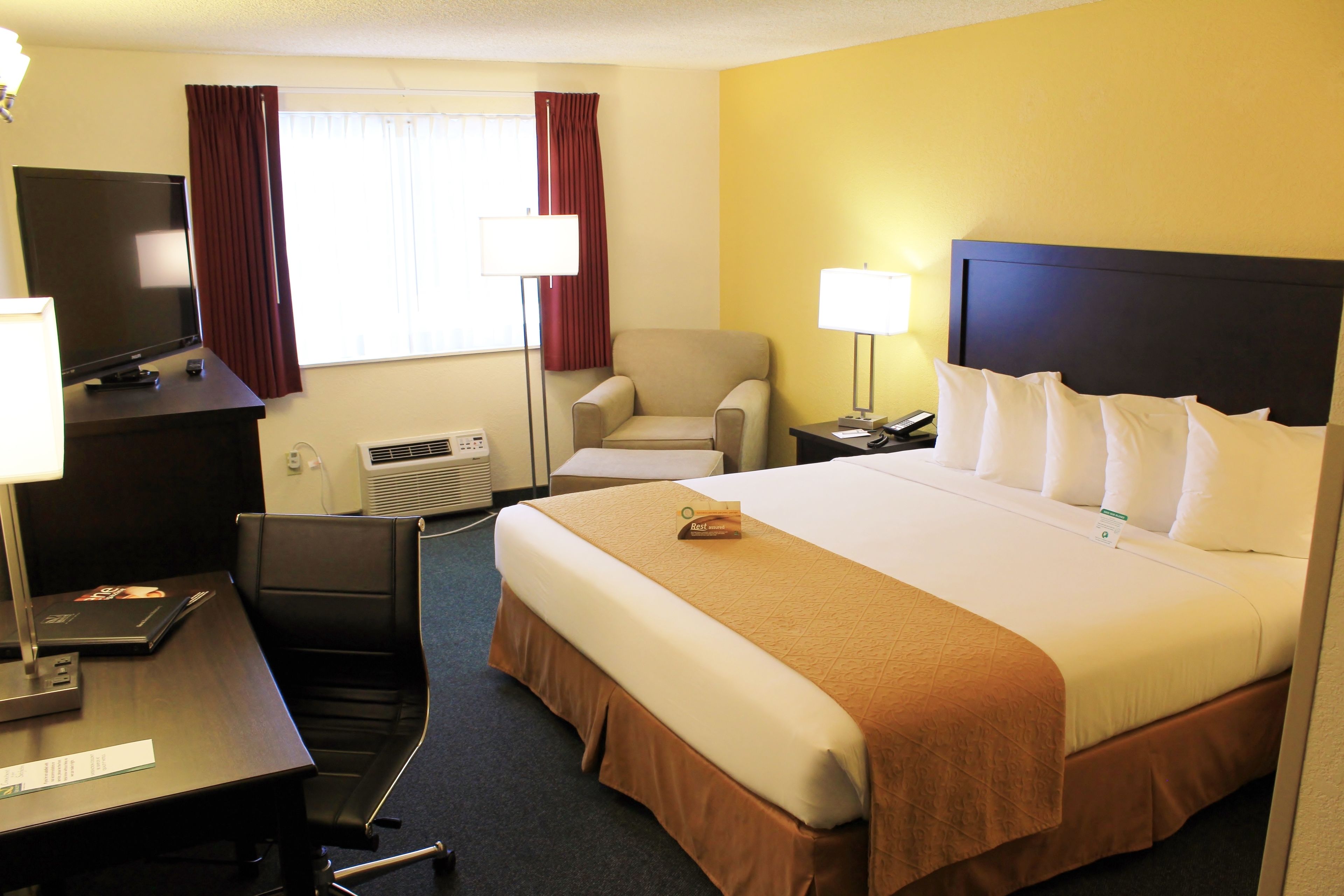 Habitación de hotel con cama, escritorio, computadora, silla, televisor y ventana con cortinas.