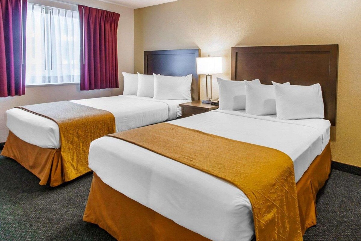 A hotel room with two beds, a nightstand, a lamp, and a window with curtains.