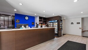 Ein Empfangsbereich mit einem hölzernen Empfangstresen, einer blauen Akzentwand, einer Uhr, einem mit Getränken gefüllten Kühlschrank und einer kleinen Pflanze.