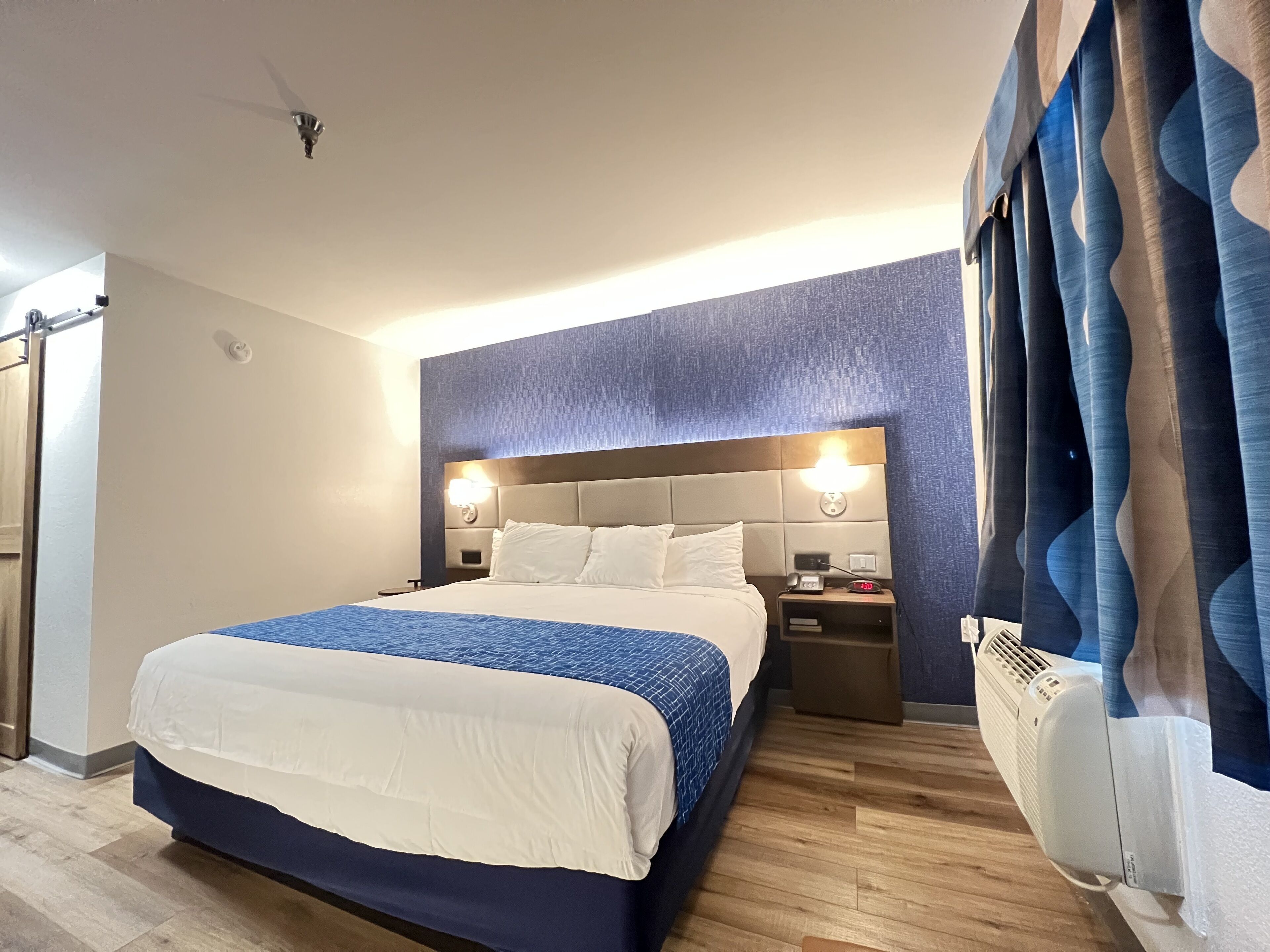 A hotel room with a large bed, two bedside tables, a wall-mounted light, and a window with curtains.