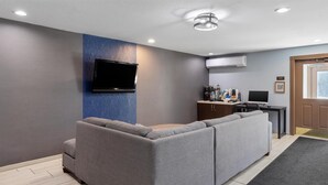 Ein modernes Wohnzimmer mit einer grauen Eckcouch, einem flachbildfernseher an der Wand, einer Kaffeemaschine und einem kleinen Schreibtisch mit einem Computer.