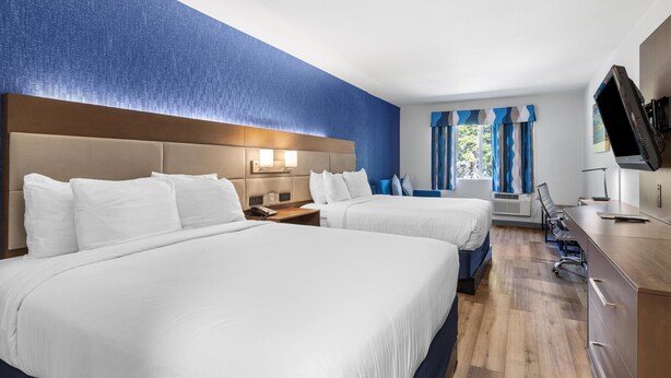 Ein Hotelzimmer mit zwei Betten, einem Schreibtisch, einem Fernseher und einem Fenster mit Vorhängen.