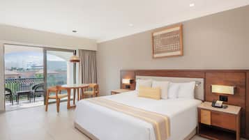 A hotel room with a large bed, a desk, and a chair, a balcony with a view of buildings, and a framed artwork on the wall.