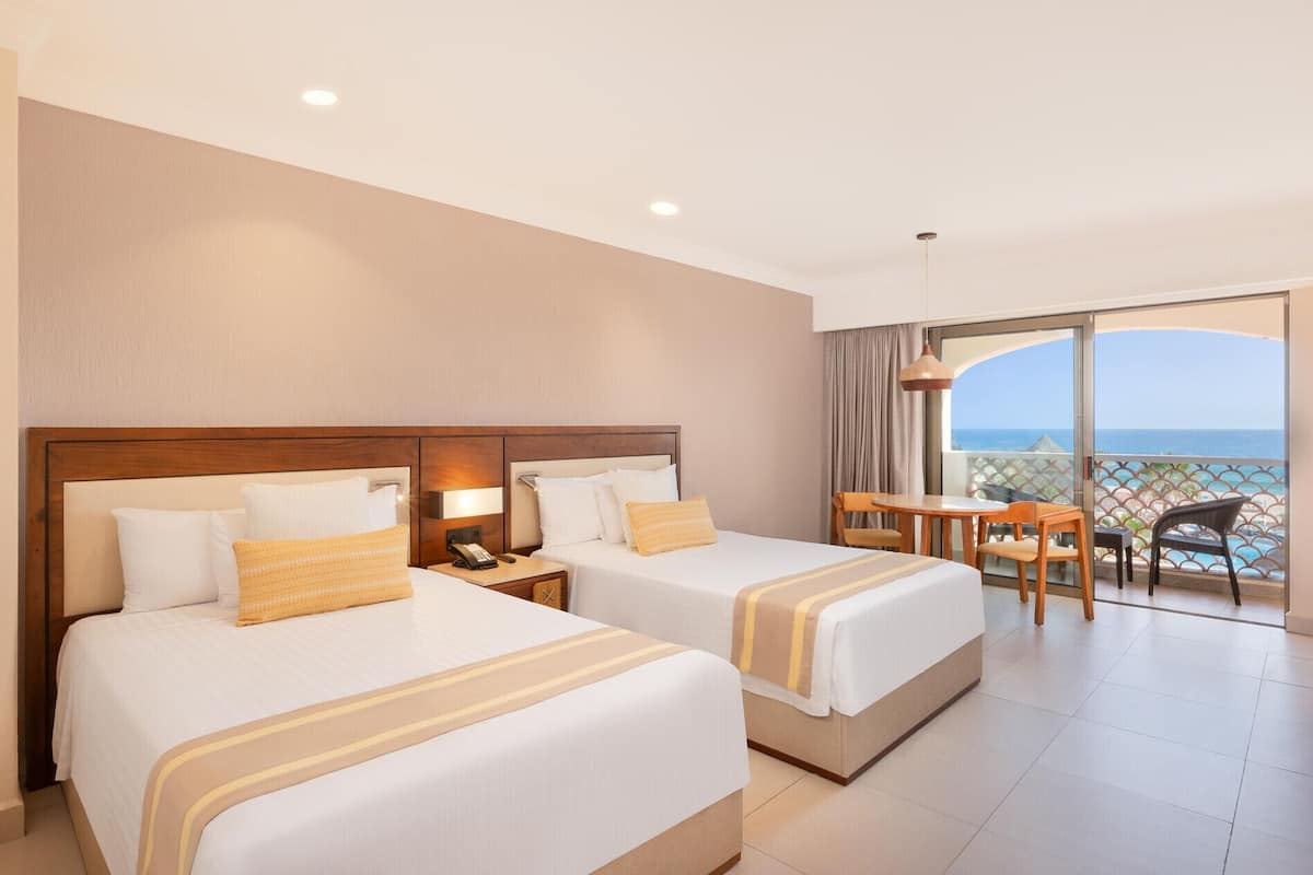 Een hotelkamer met twee bedden, een kleintje tafeltje en een balkon met uitzicht op zee.