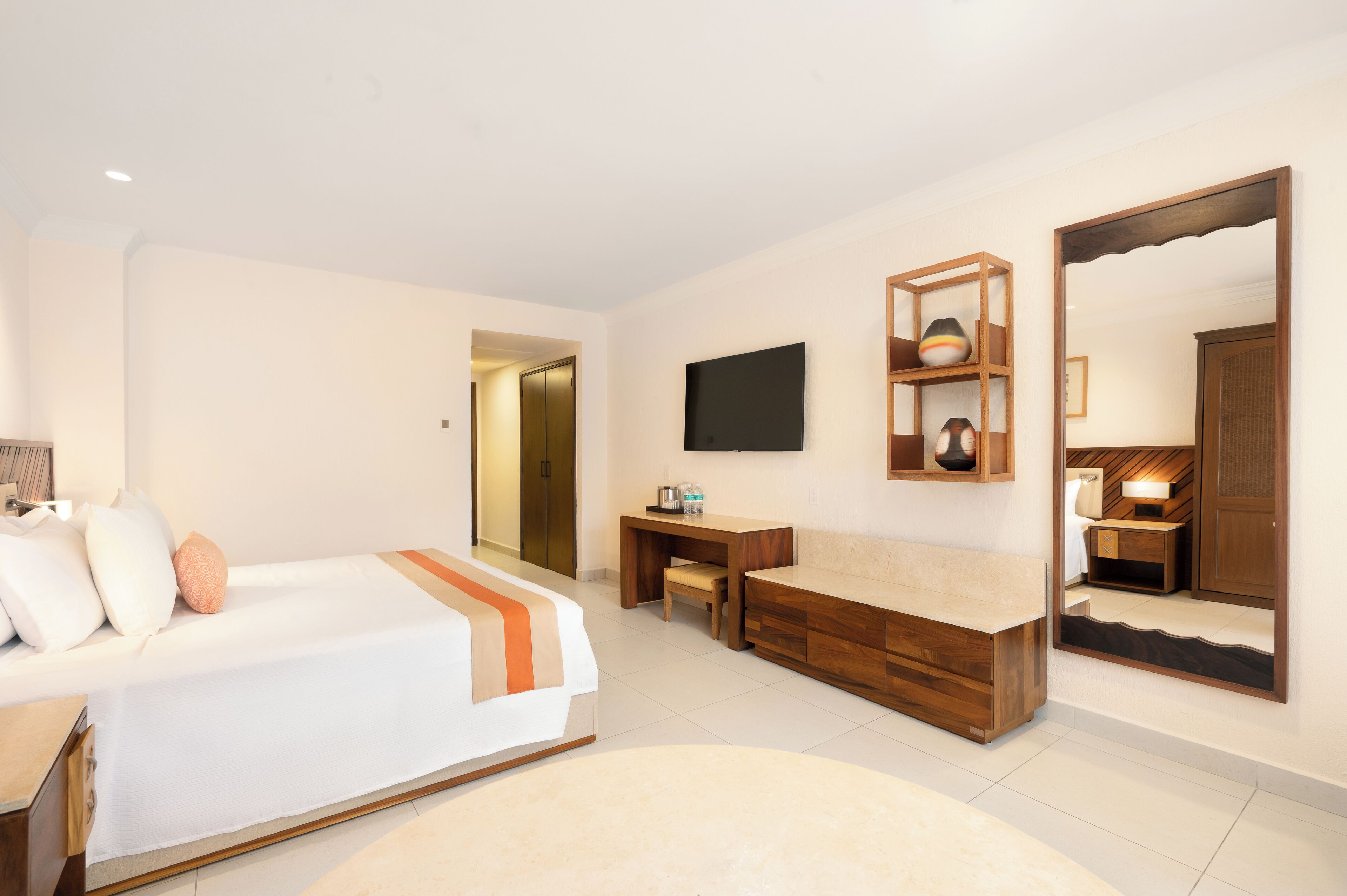 Ein modernes Hotelzimmer mit einem Bett, einer hölzernen Sitzbank, einem kleinen Tisch, einem Fernseher und einem großen Spiegel.