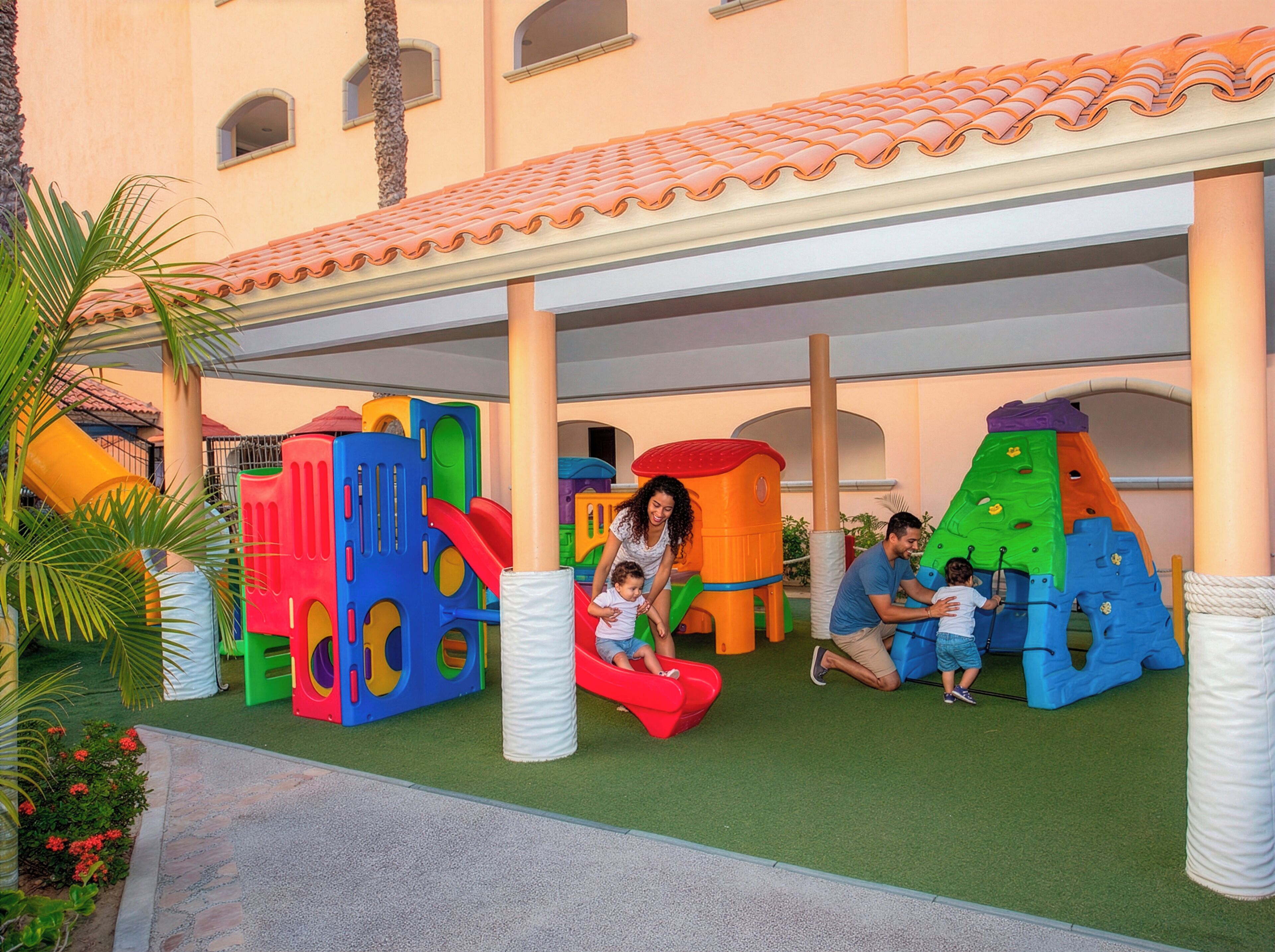 Un área de juegos infantiles con estructuras coloridas, un espacio de descanso techado y un edificio con tejado de baldosas.