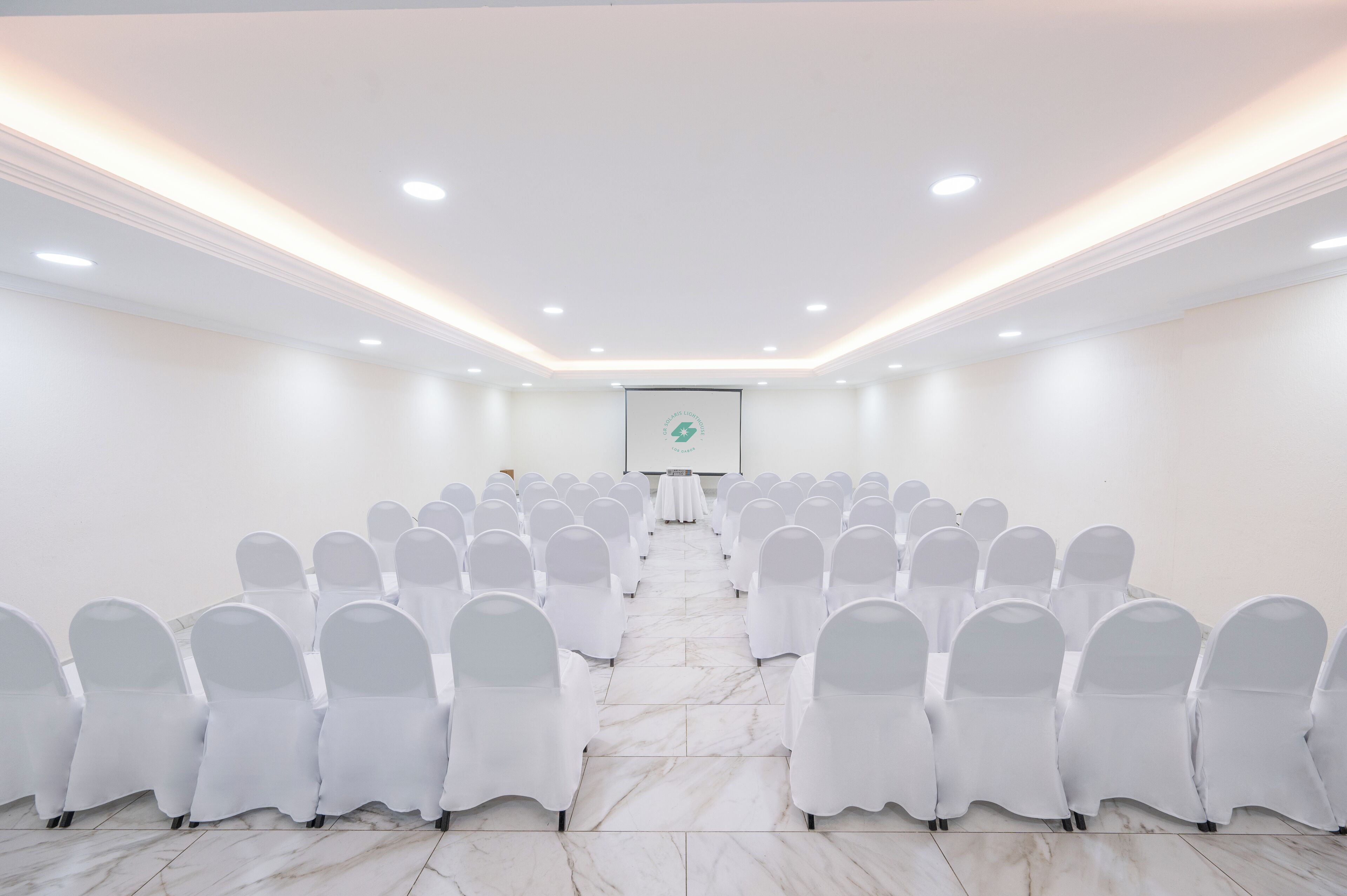 Un salón de conferencias con filas de sillas blancas, una pantalla de proyector al frente y piso de mármol.