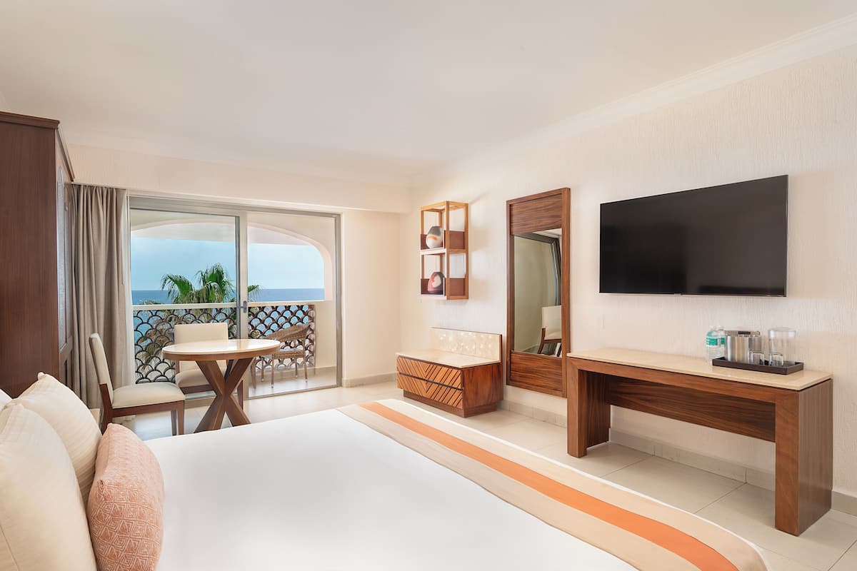 Een hotelkamer met een bed, een bureau, een televisie, een balkon met een tafel en stoelen, en uitzicht op de oceaan.