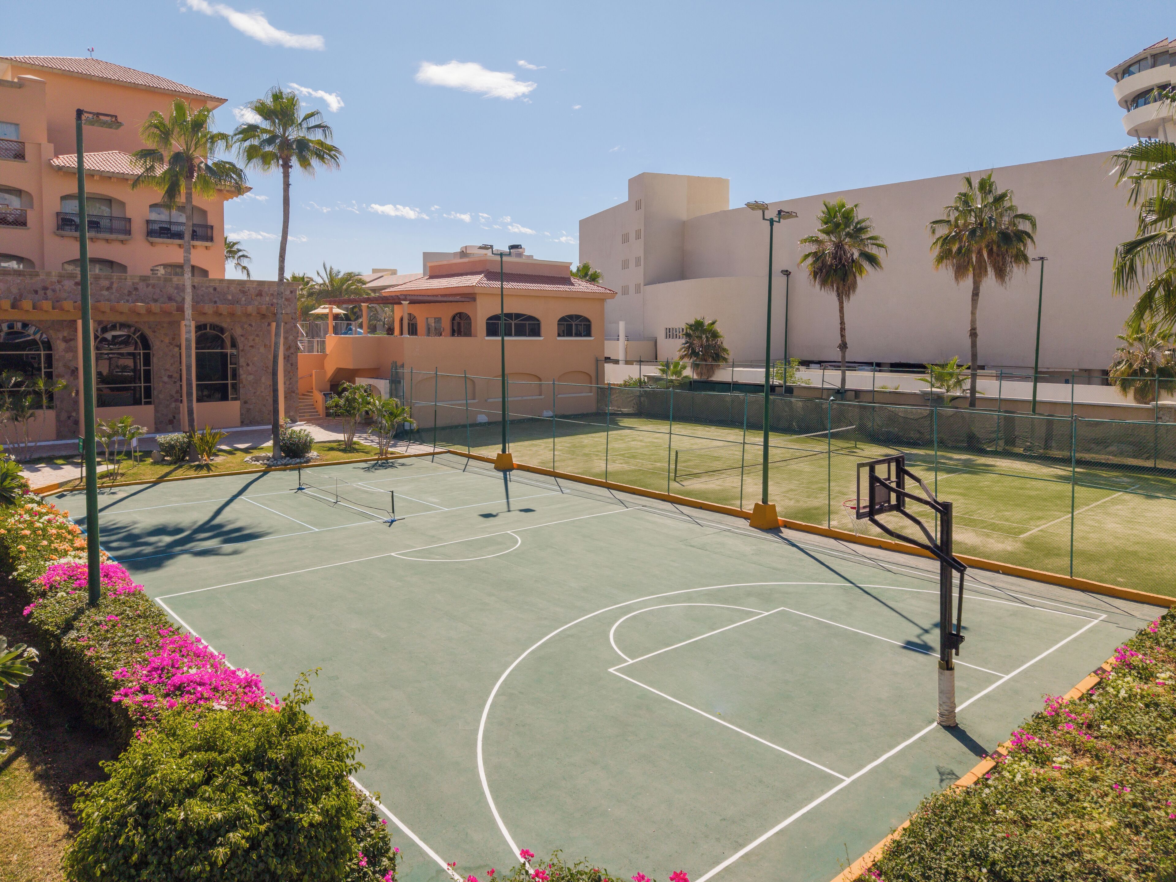 An outdoor basketball court with a green surface, surrounded by a fence, and adjacent to a building with a beige facade and arched windows.