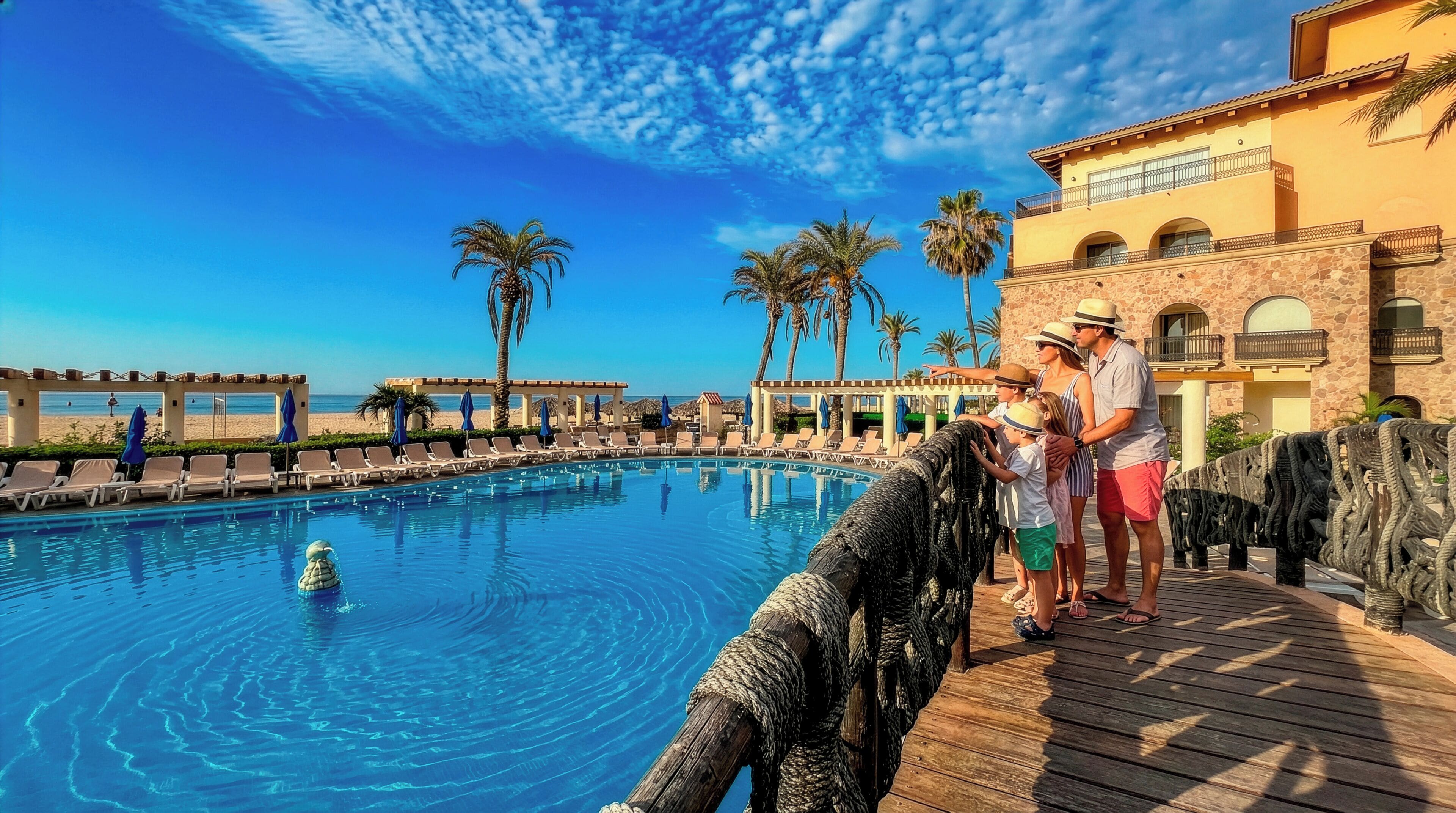A pool with a wooden deck, palm trees, and a building with arched windows.