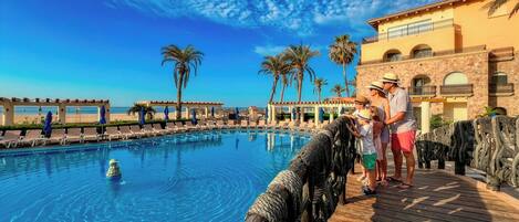 A pool with a wooden deck, palm trees, and a building with arched windows.