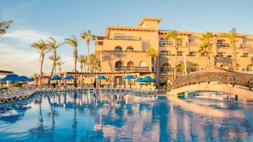 Relax by the resort's large pool surrounded by lounge chairs and umbrellas, with a multi-story building and arched bridge nearby.