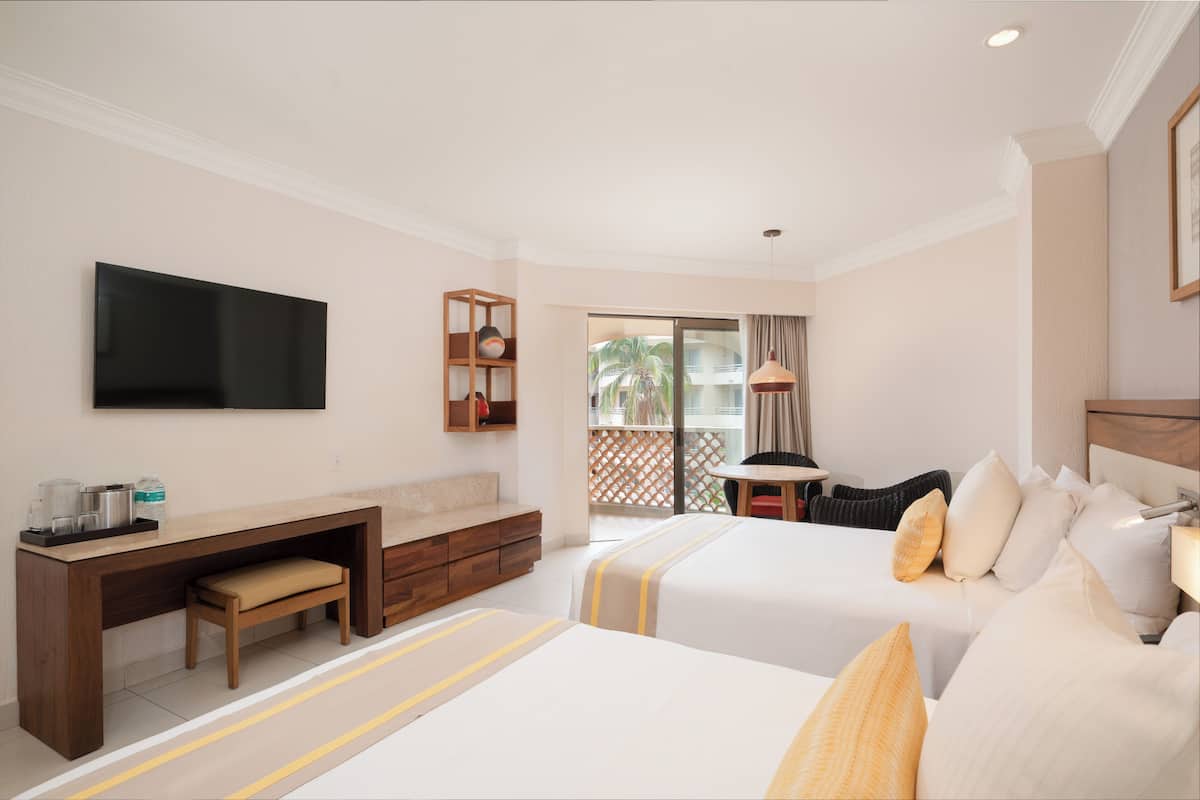 Een hotelkamer met twee bedden, een bureau, een televisie en een balkon met uitzicht op palmbomen.