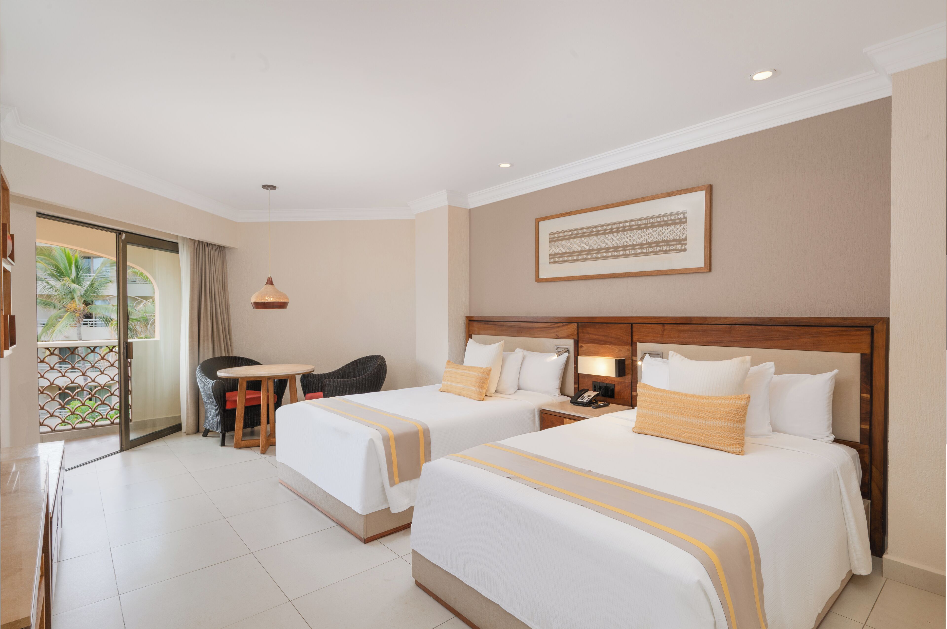 A hotel room with two beds, a small table, a chair, and a balcony with a view of palm trees.