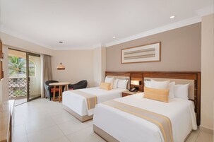 Habitación de hotel con dos camas, una mesita, una silla y un balcón con vista a palmeras.