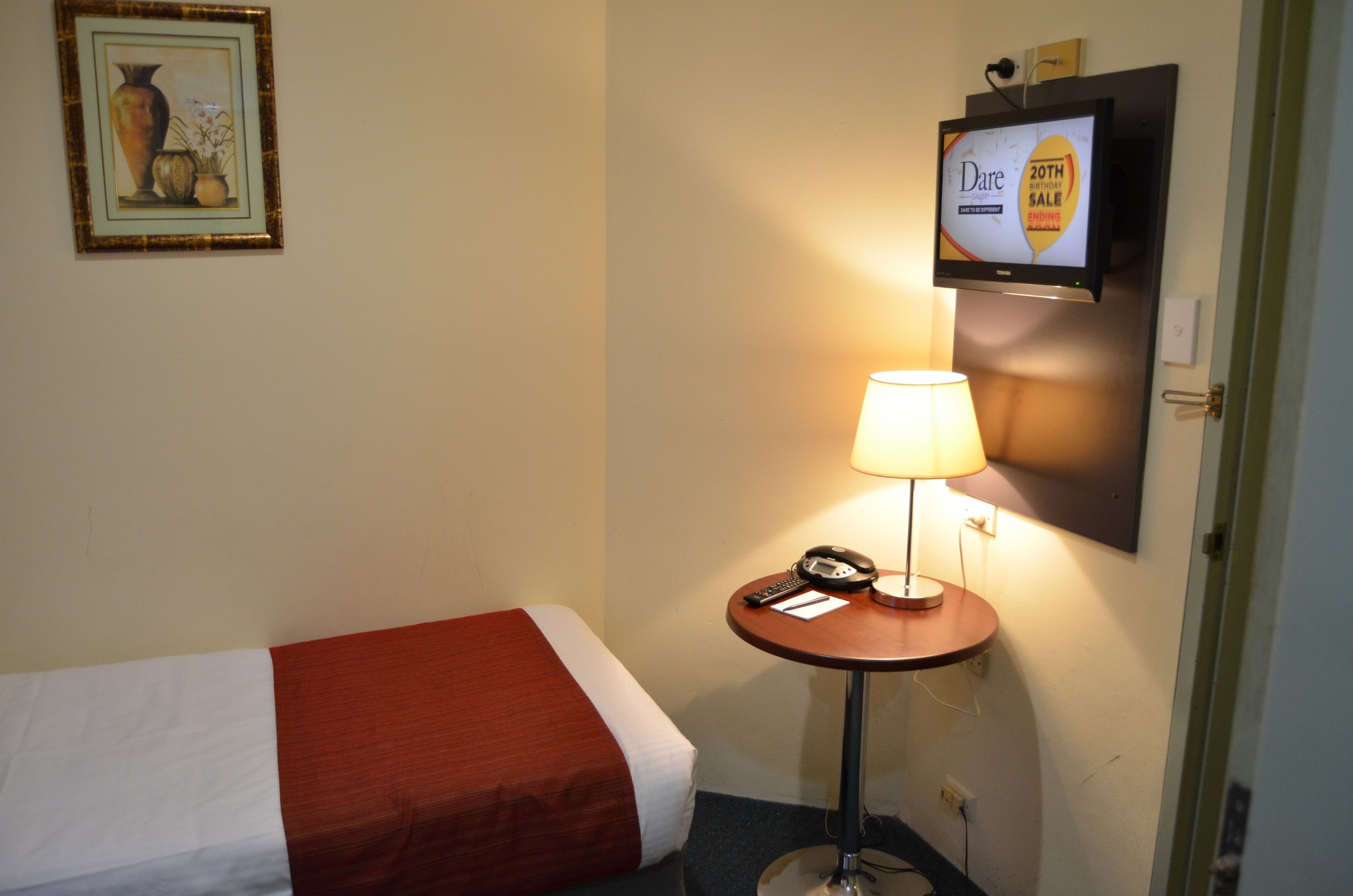 A hotel room with a bed, a bedside table with a lamp, a TV mounted on the wall, and a framed picture on the wall.