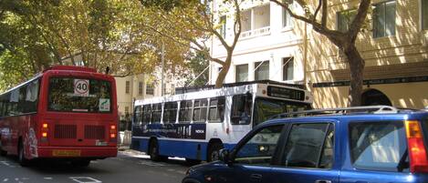 Vista urbana con un autobús rojo, un coche azul y un autobús blanco. Hay árboles y un edificio con un letrero que dice 'Central Railway St.'