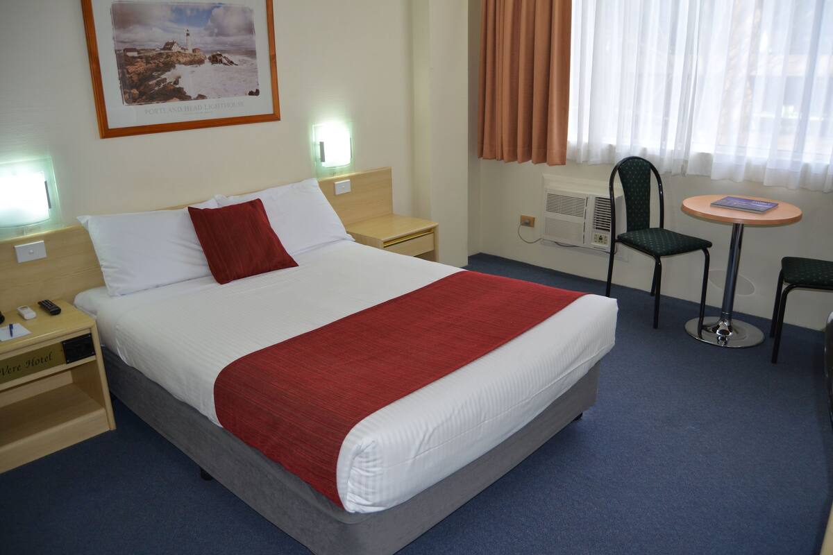 A hotel room with a bed, a nightstand, a chair, a small table, and a wall-mounted air conditioner.