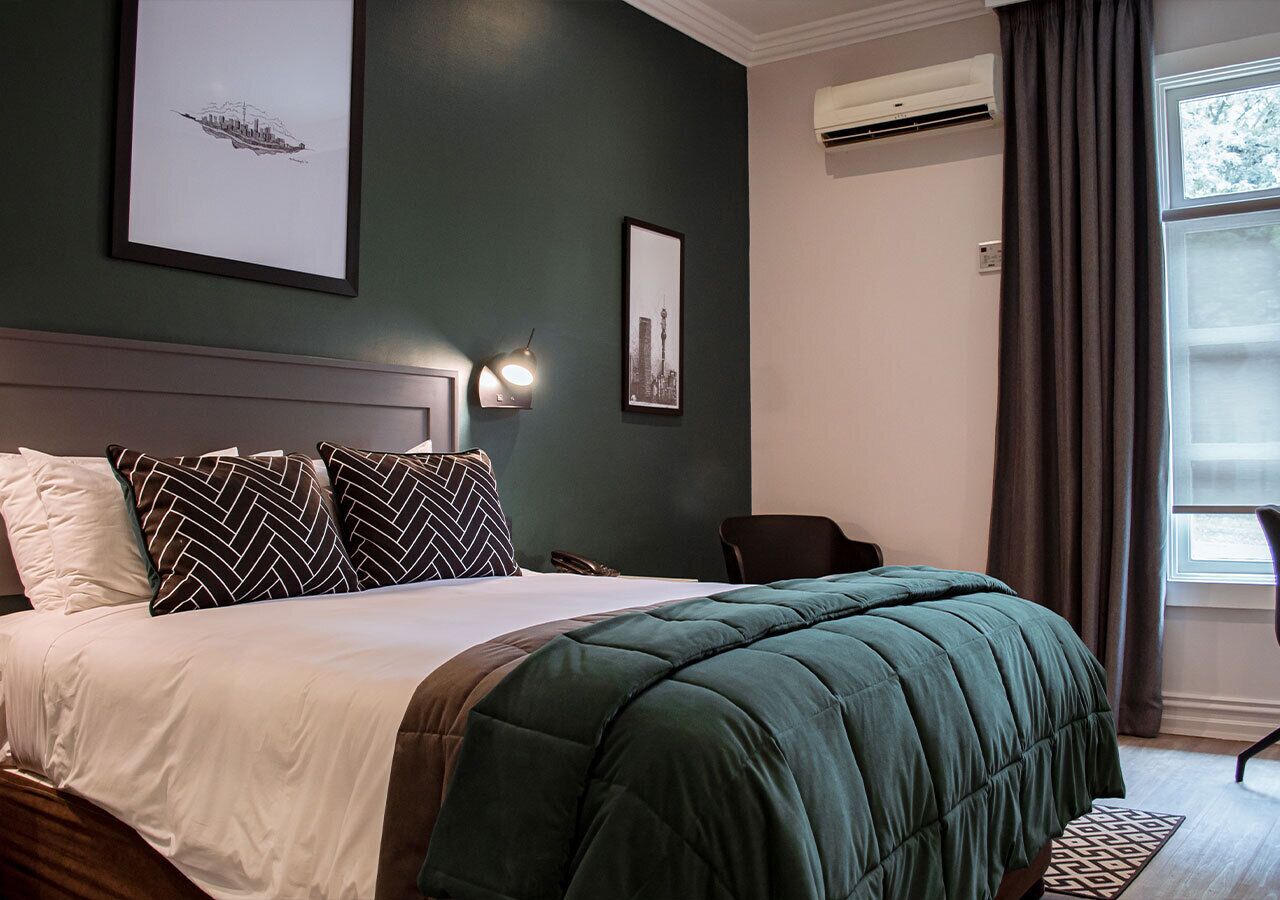 Una cama bien tendida con una manta verde y almohadas estampadas, una lámpara de noche y cuadros en la pared.