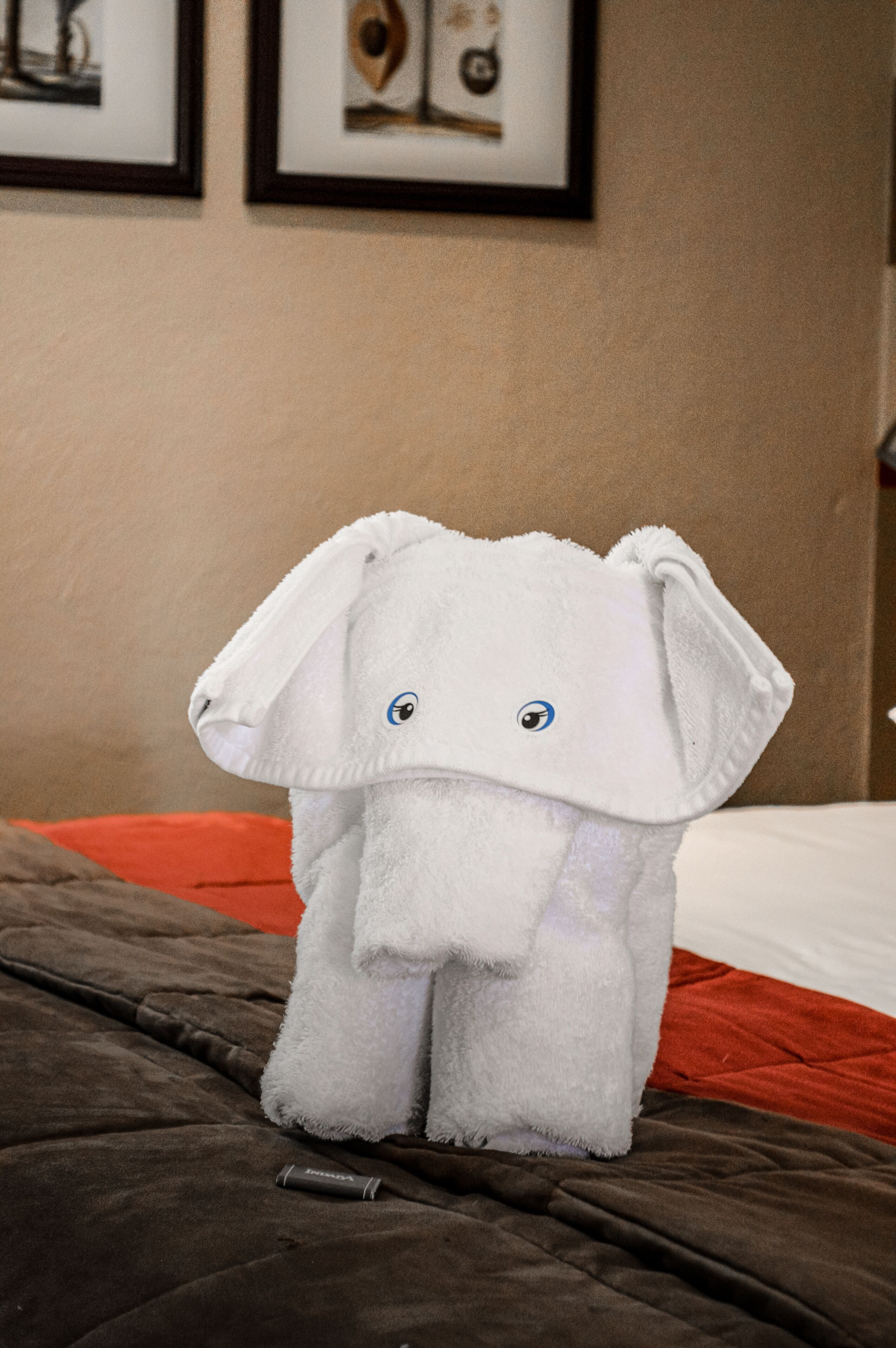 Uma toalha macia em forma de elefante sobre uma cama com cobertura marrom e almofada laranja.