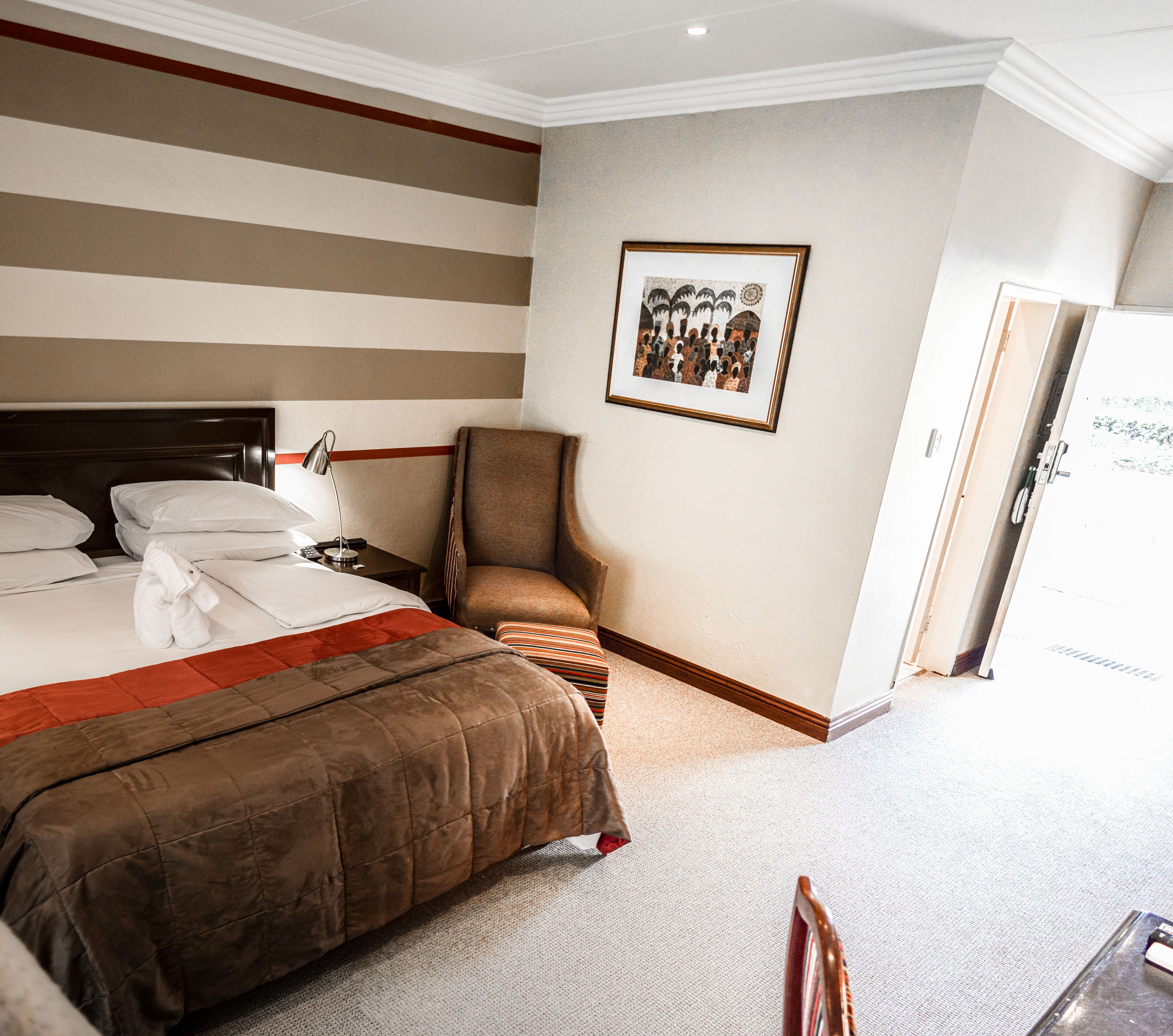 A hotel room with a large bed, a chair, a desk with a lamp, and a framed picture on the wall.