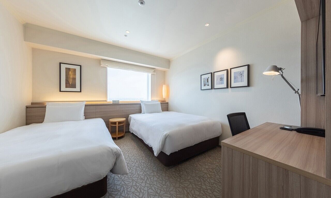 A hotel room with two beds, a desk, a chair, a lamp, and framed pictures on the wall.