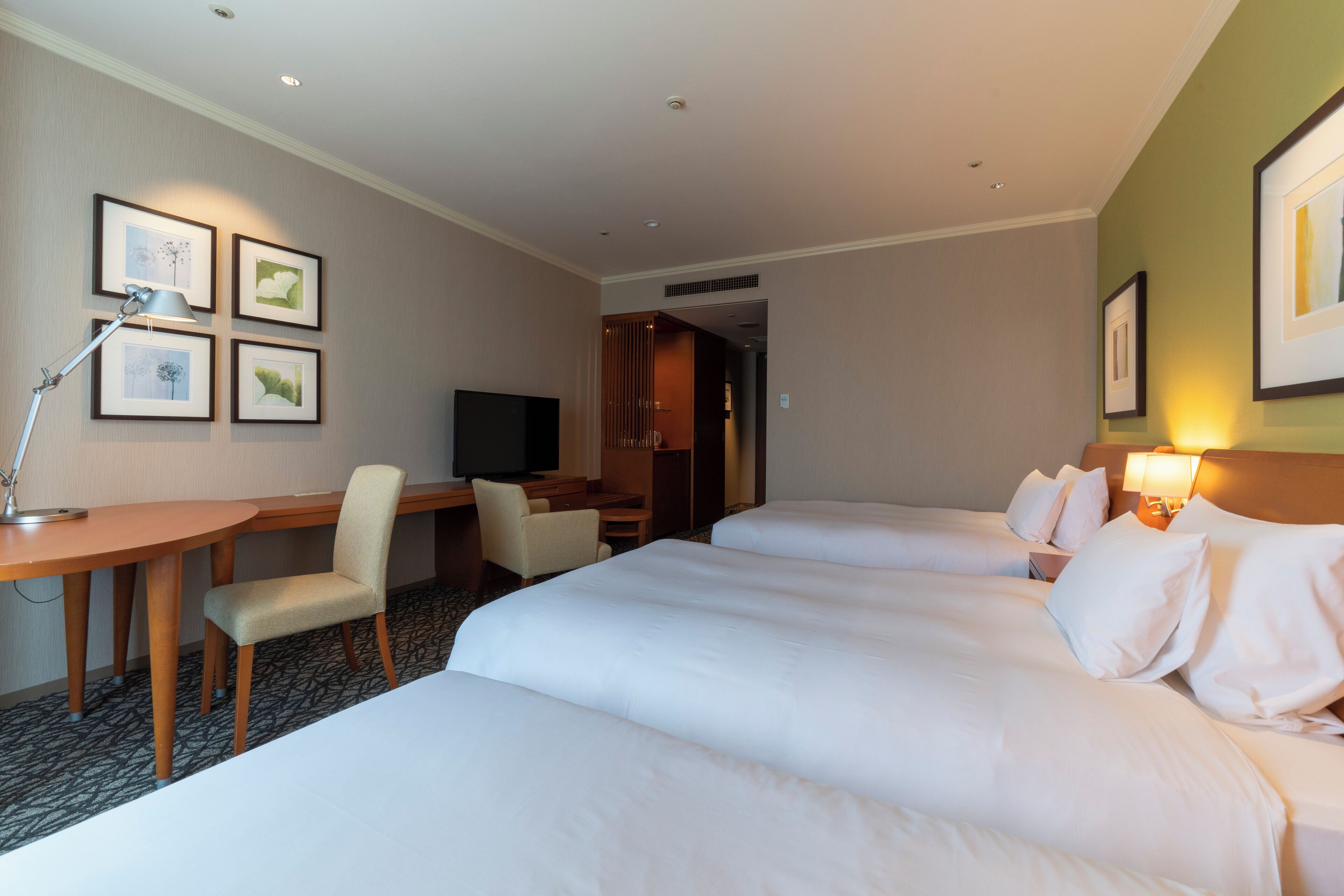 A hotel room with two beds, a desk, a chair, a TV, and framed pictures on the wall.