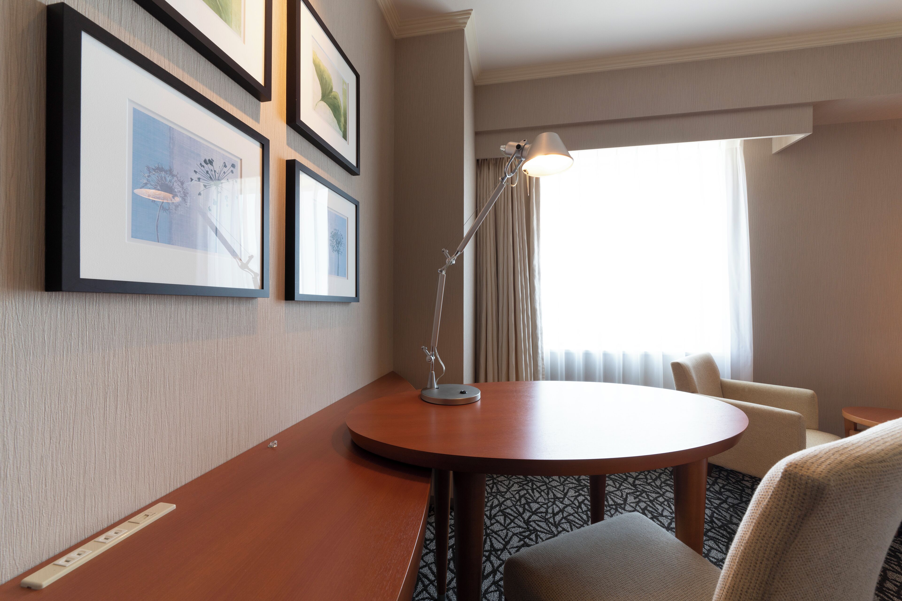 A hotel room with a round table, a lamp, a chair, and framed pictures on the wall.