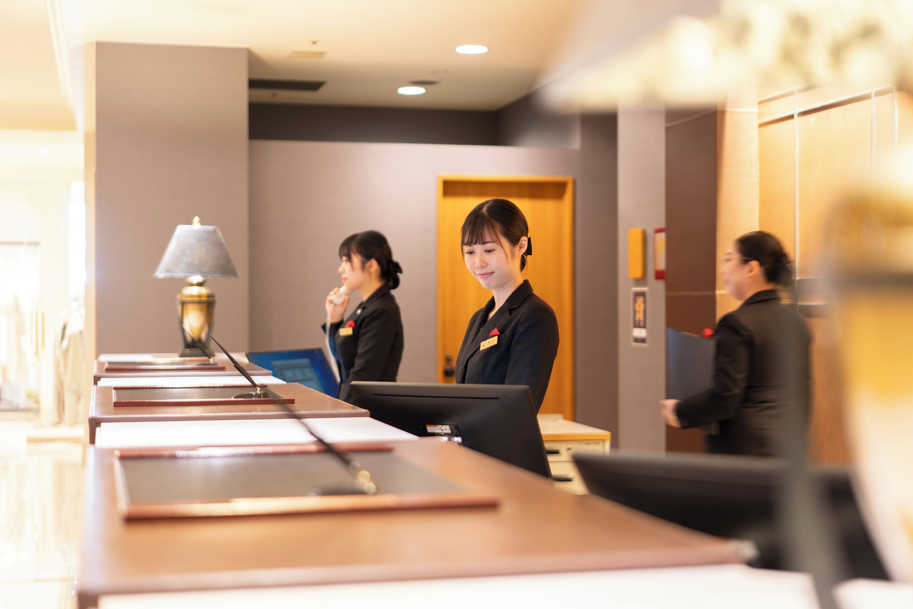 A reception desk in a hotel lobby with a lamp, a computer, and a wooden door in the background.