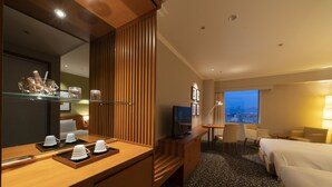 A hotel room with a bed, a desk with a computer, a TV, a chair, a sofa, and a view of the city at dusk. - Hotel Associa Toyohashi (Toyohashi)