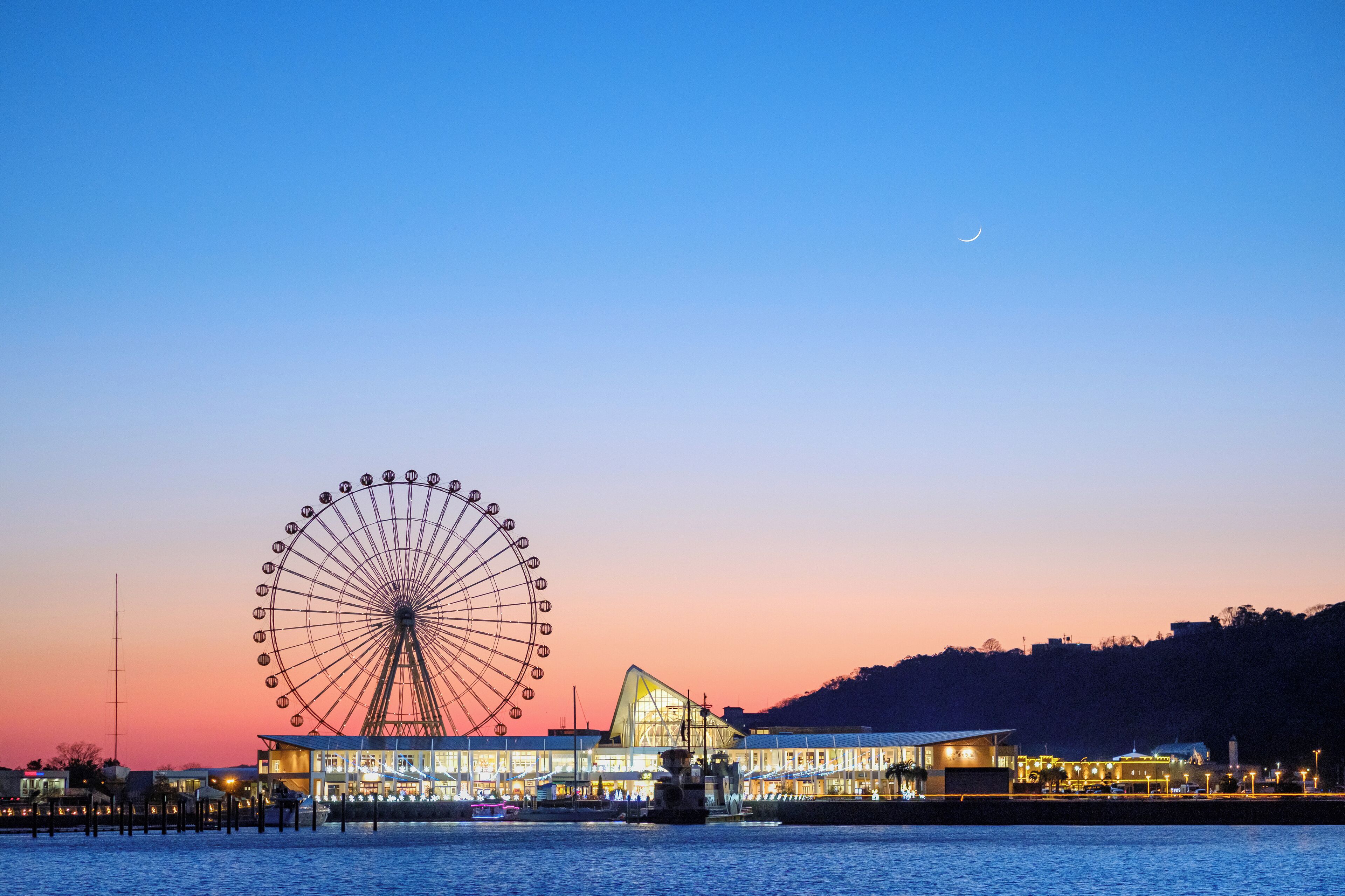 A waterfront view with a large Ferris wheel and a modern building with a triangular roof.