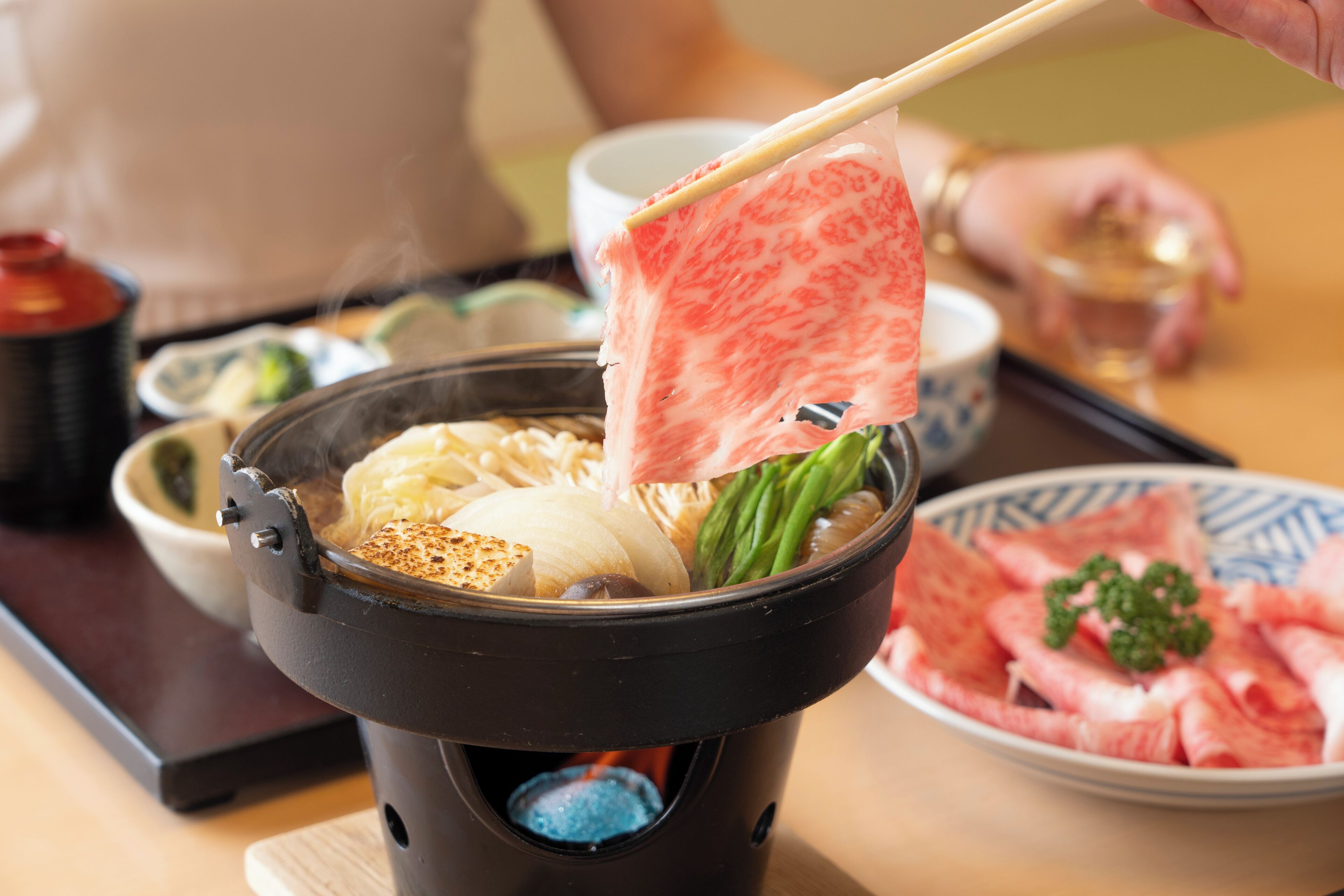 Una olla caliente con diversos ingredientes, un tazón de carne en rodajas y una taza de té.