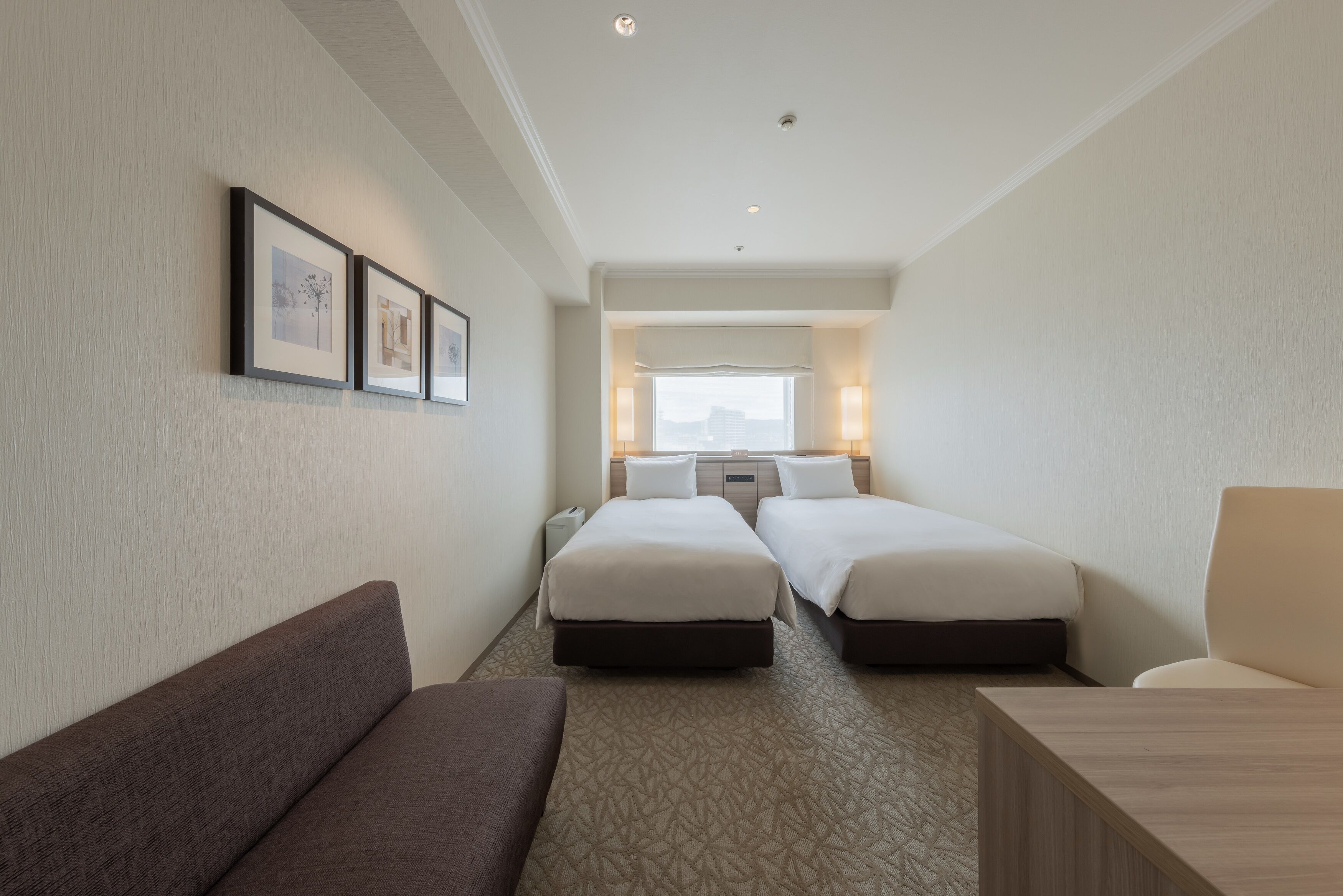 A hotel room with two beds, a sofa, a desk, and framed pictures on the wall.