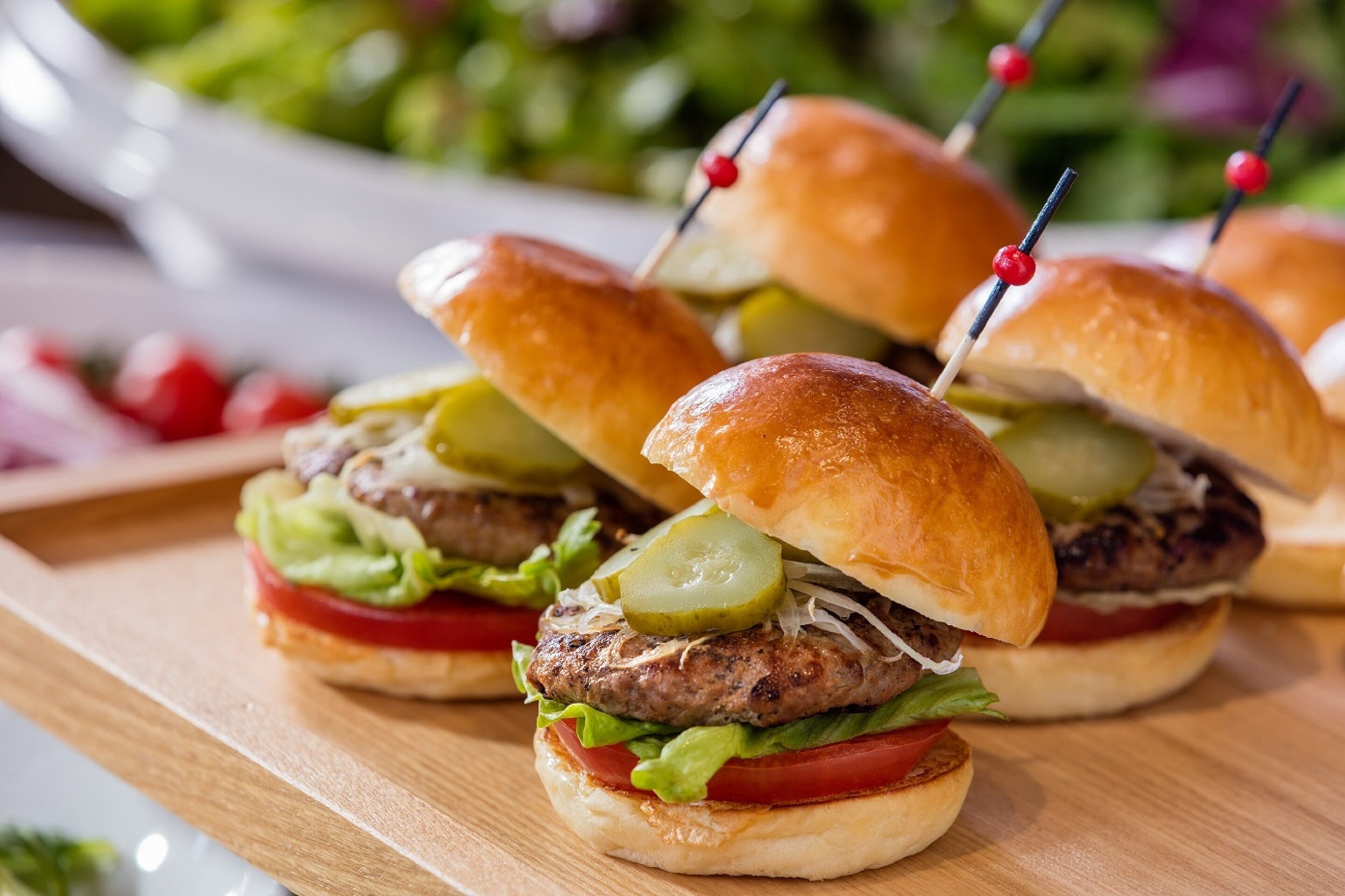 Three burgers with pickles and toothpicks on buns, placed on a wooden board.