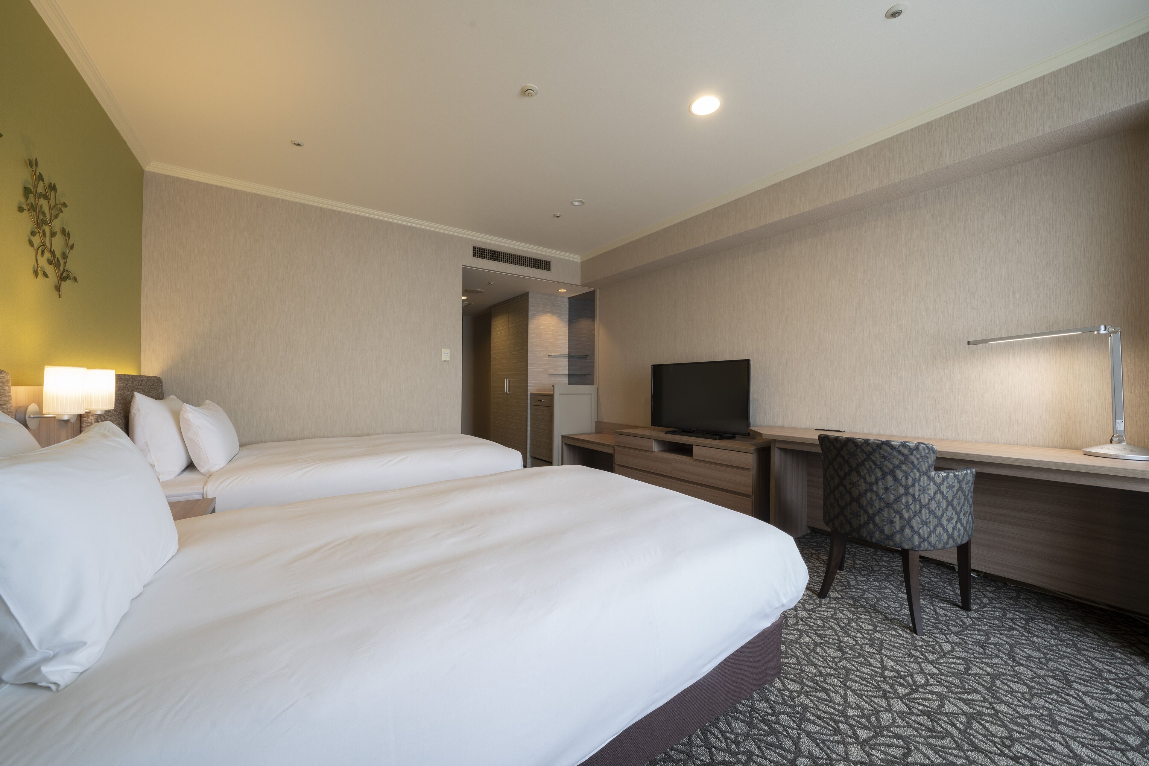 A hotel room with two beds, a desk, a chair, a television, and a lamp.