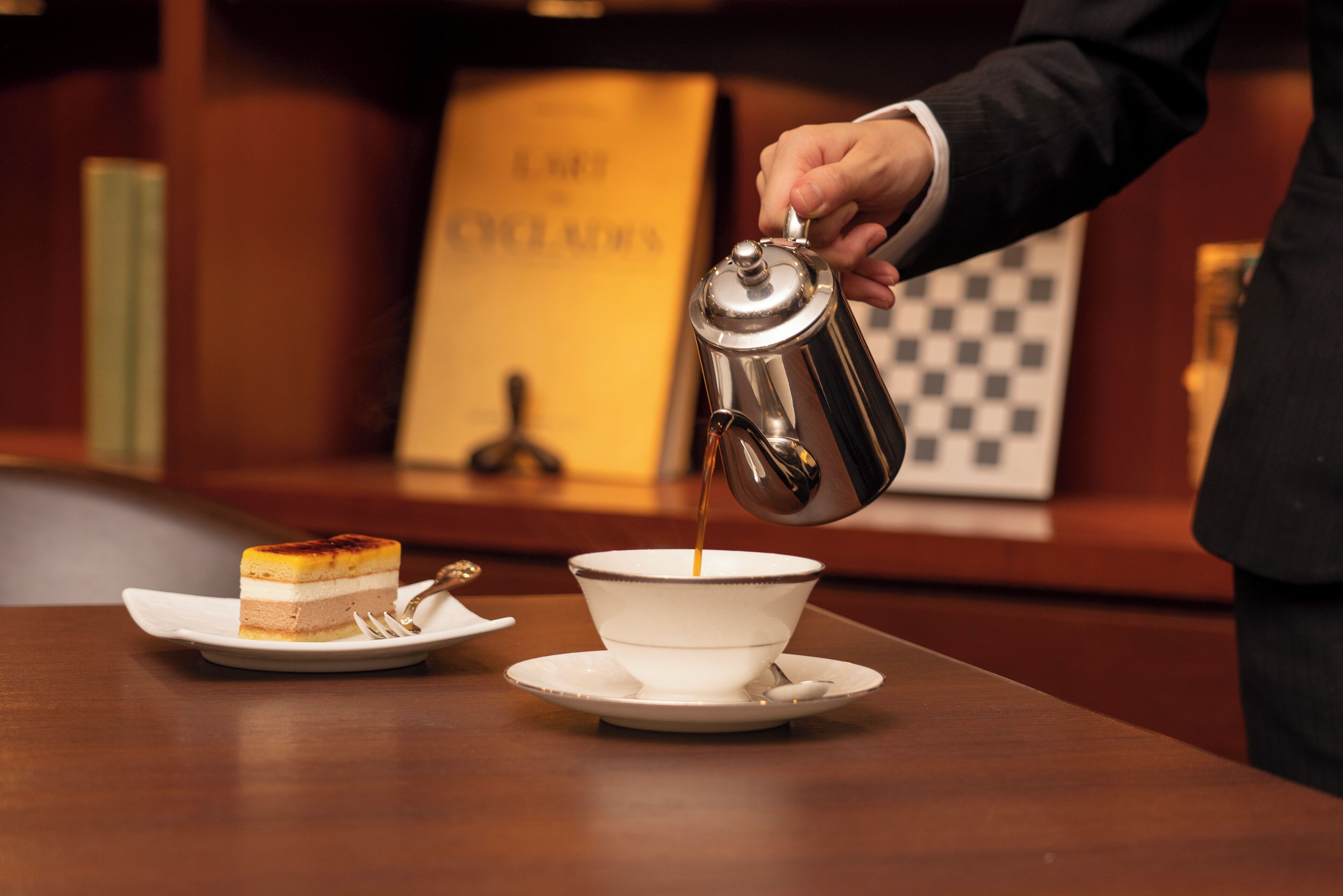 A person pouring coffee into a cup on a table with a slice of cake and a spoon.