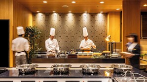 A modern kitchen with chefs preparing food, stainless steel cooking stations, and a decorative wall with a leaf pattern. - Hotel Associa Toyohashi (Toyohashi)
