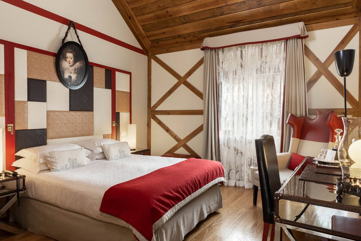 A hotel room with a large bed, a wooden ceiling, a wall-mounted picture, a dining table with chairs, and a window with curtains.