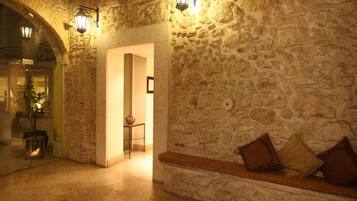 A stone-walled hallway with a bench and cushions, a door leading to another room, and two wall-mounted lanterns.