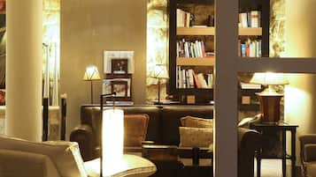 Una sala de estar acogedora con un sofá de cuero, una mesita de centro y una estantería llena de libros. Dos lámparas de pie proporcionan luz.